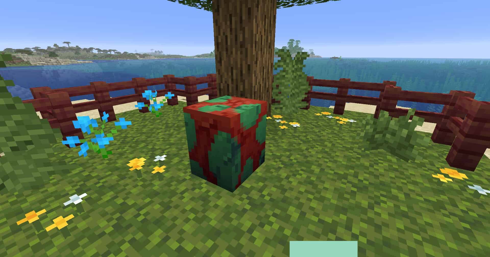 Zrzut ekranu z gry Minecraft, przedstawiający częściowo pęknięte jajo niuchacza w ogródku otoczonym płotem. Jajo jest koloru zielono-czerwonego i ma kształt sześcienny. W ogródku na blokach mchu widzimy żółte, białe i niebieskie kwiatki, oraz parę zielonych paproci. Za jajem widać konar drzewa dębowego i nieco liści. Ogródek otacza płot z drewna namorzynowego o ciemnoczerwonej barwie. W tle w oddali widzimy ocean i niebo. 