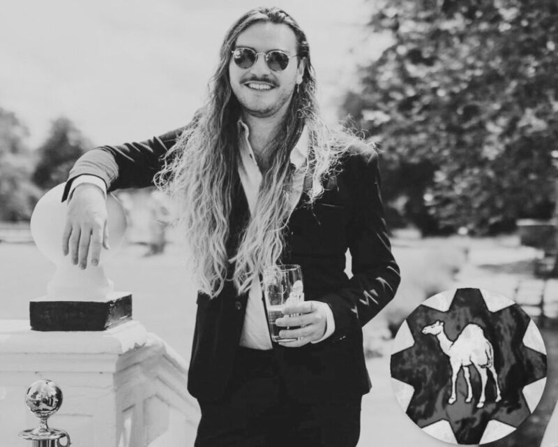 Czarno-białe zdjęcie przedstawiające YouTubera Camelworksa. Mężczyzna w średnim wieku, z długimi włosami, okularami, ubrany w garnitur stoi na tle parku opierając się o murek.