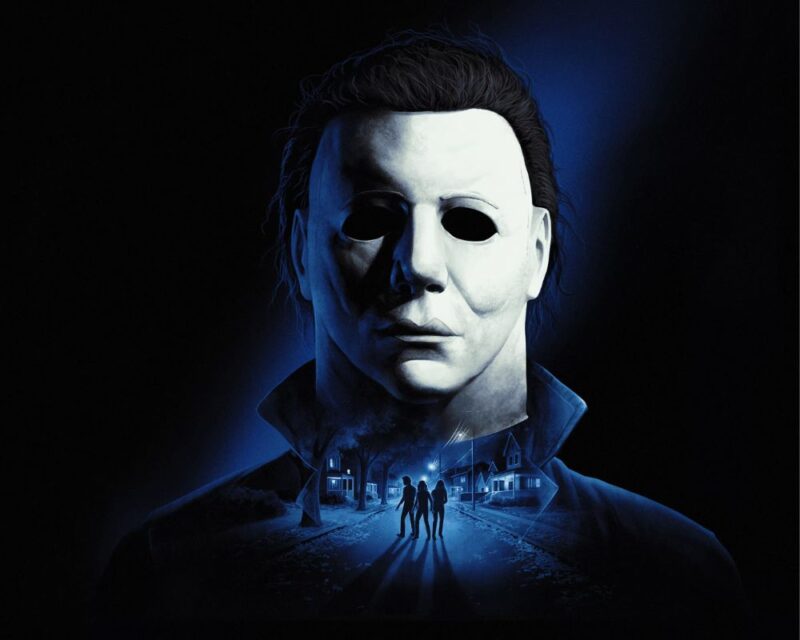 Mroczny plakat gry Halloween ukazujący zamaskowanego zabójcę w charakterystycznej białej masce patrzącego prosto przed siebie, a poniżej nocną ulicę z trzema osobami uciekającymi między domami na przedmieściach.