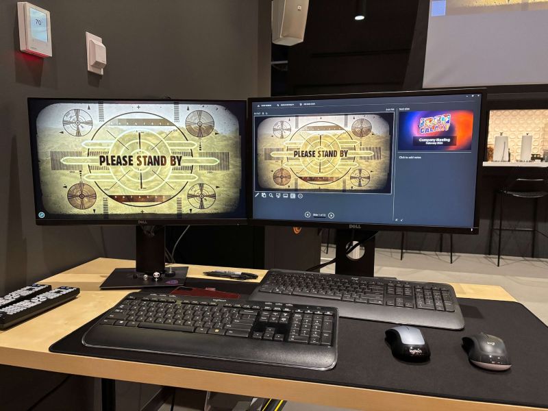 Nowoczesne stanowisko pracy z dwoma monitorami Dell, klawiaturą i myszą na biurku, na ekranach grafika „Please Stand By” w stylu retro z gry Fallout. Kolorystyka zdjęcia jest stonowana i nowoczesna, z przewagą ciemnych oraz neutralnych barw. Dominują odcienie czerni, grafitu i szarości widoczne w monitorach, klawiaturach, myszach oraz ścianach w tle. Biurko w jasnym, naturalnym kolorze drewna.