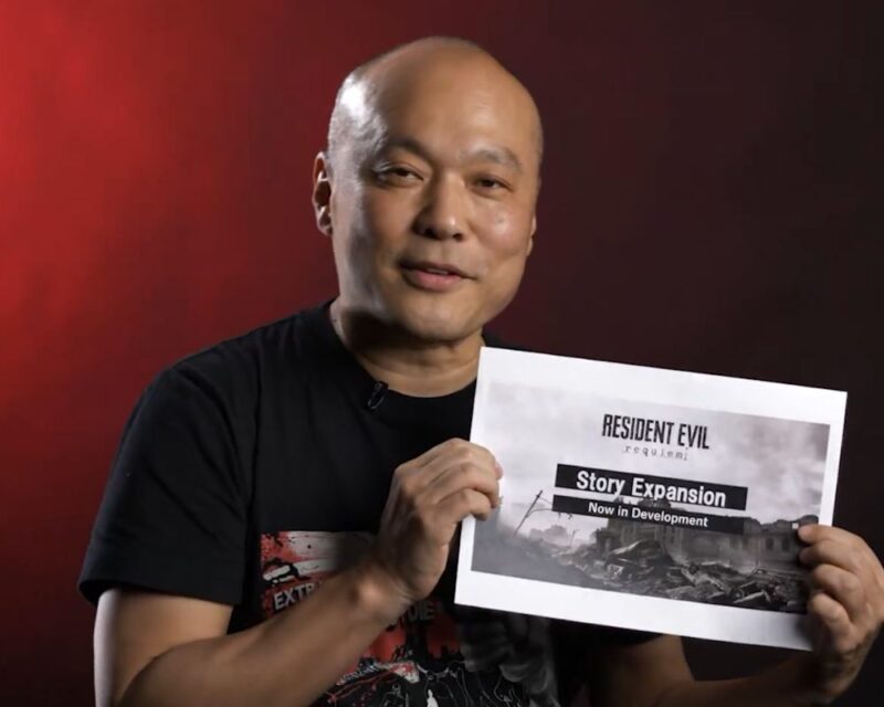 Uśmiechnięty, łysy mężczyzna w czarnej koszulce siedzi na tle czerwonego, studyjnego oświetlenia i trzyma w obu dłoniach wydrukowaną kartkę. Na kartce widnieje napis „Resident Evil Requiem” oraz „Story Expansion – Now in Development”, a poniżej grafika przedstawiająca zrujnowane miasto. Mężczyzna prezentuje kartkę w stronę kamery, jak podczas zapowiedzi lub ogłoszenia dotyczącego gry.