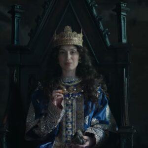 Stopklatka ze zwiastuna ujawniającego prace nad Total War: Medieval 3. Na obrazie widzimy postać władczyni w koronie, przypuszczalnie więc królową, odzianą w niebieskie szaty z bogatymi zdobieniami ze złota i pereł. Siedzi na drewnianym tronie. Ciemne, kręcone włosy ma rozpuszczone. W dłoniach trzyma monetę i mieszek.