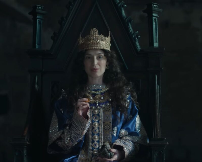 Stopklatka ze zwiastuna ujawniającego prace nad Total War: Medieval 3. Na obrazie widzimy postać władczyni w koronie, przypuszczalnie więc królową, odzianą w niebieskie szaty z bogatymi zdobieniami ze złota i pereł. Siedzi na drewnianym tronie. Ciemne, kręcone włosy ma rozpuszczone. W dłoniach trzyma monetę i mieszek.