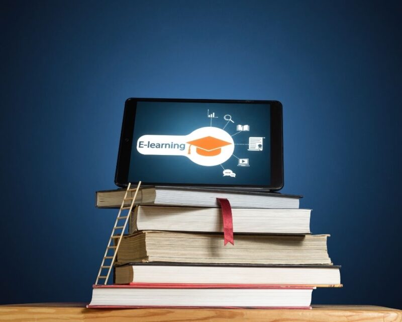 Książki ustawione na granatowym tle i drewnianym blacie, na książkach postawiony jest laptop z napisem e-learnig z czapką uniwersytecką oraz ikonami związanymi z dziedzinami nauki