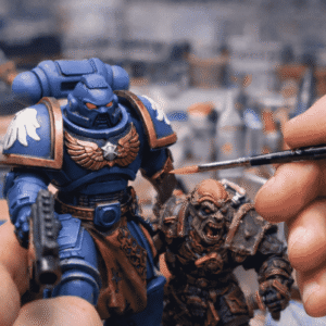 Zbliżenie na ręcznie malowane figurki z uniwersum Warhammera – potężnego Space Marine’a w niebieskim pancerzu oraz zielonoskórego Orka. W kadrze widoczna jest dłoń z precyzyjnym pędzlem, nanosząca detale farbą, co podkreśla rzemieślniczy charakter hobby i dbałość o każdy szczegół. Obraz oddaje pasję, skupienie i kunszt, które sprawiają, że figurki Warhammera żyją także poza ekranem komputera