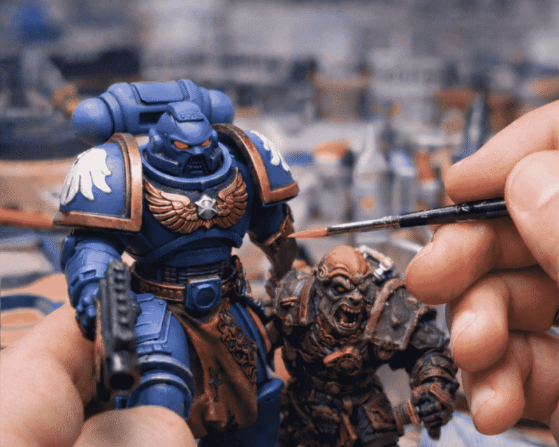 Zbliżenie na ręcznie malowane figurki z uniwersum Warhammera – potężnego Space Marine’a w niebieskim pancerzu oraz zielonoskórego Orka. W kadrze widoczna jest dłoń z precyzyjnym pędzlem, nanosząca detale farbą, co podkreśla rzemieślniczy charakter hobby i dbałość o każdy szczegół. Obraz oddaje pasję, skupienie i kunszt, które sprawiają, że figurki Warhammera żyją także poza ekranem komputera