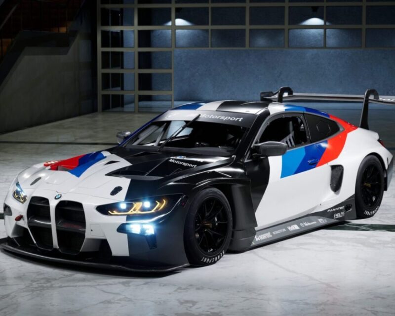 Wyścigowe BMW M4 GT3 w barwach BMW Motorsport (biało-niebiesko-czerwonych) stoi w nowoczesnym, oświetlonym garażu. Samochód ma agresywny pakiet aerodynamiczny z dużym przednim splitterem, poszerzonymi nadkolami i wysokim tylnym skrzydłem. Przednie reflektory świecą na niebiesko, a nadwozie łączy białe, czarne i kolorowe akcenty charakterystyczne dla działu Motorsport.