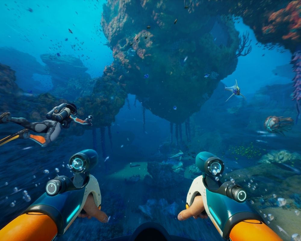 Zrzut ekranu z gry Subnautica 2 przedstawiający podwodną scenę widzianą z perspektywy pierwszej osoby. Na pierwszym planie widoczne są ręce postaci w pomarańczowym skafandrze nurkowym, na których zamontowane są futurystyczne urządzenia przypominające rękawice lub narzędzia z wbudowanymi lampami. Wokół unoszą się pęcherzyki powietrza. Po lewej stronie płynie inny nurek w jasnym skafandrze z plecakiem tlenowym, wyciągający rękę przed siebie. W centrum kadru znajduje się ogromna formacja skalna porośnięta koralowcami i roślinnością, unosząca się nad dnem oceanu, z której zwisają długie, przypominające korzenie struktury. W wodzie pływają liczne ryby i morskie stworzenia, a w tle widać dalsze fragmenty podwodnego krajobrazu z rafami i skałami skąpanymi w jasnoniebieskim świetle oceanu.
