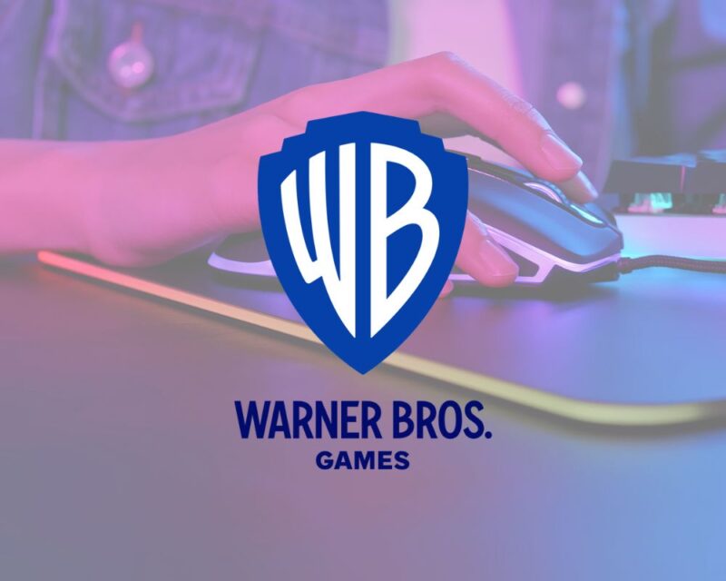 Logotyp Warner Bros. Games nałożony na zdjęcie dłoni gracza na myszce gamingowej. Logo to tarcza z literami 'WB' (białymi na niebieskim tle), pod którą znajduje się napis 'WARNER BROS. GAMES' w kolorze ciemnoniebieskim. Tło pokazuje dłoń w zbliżeniu na myszce z podświetleniem LED, na podkładce, w nastrojowym różowo-niebieskim świetle. Ilustracja sugeruje zwolnienia w Warner Bros. Montreal w 2026 roku