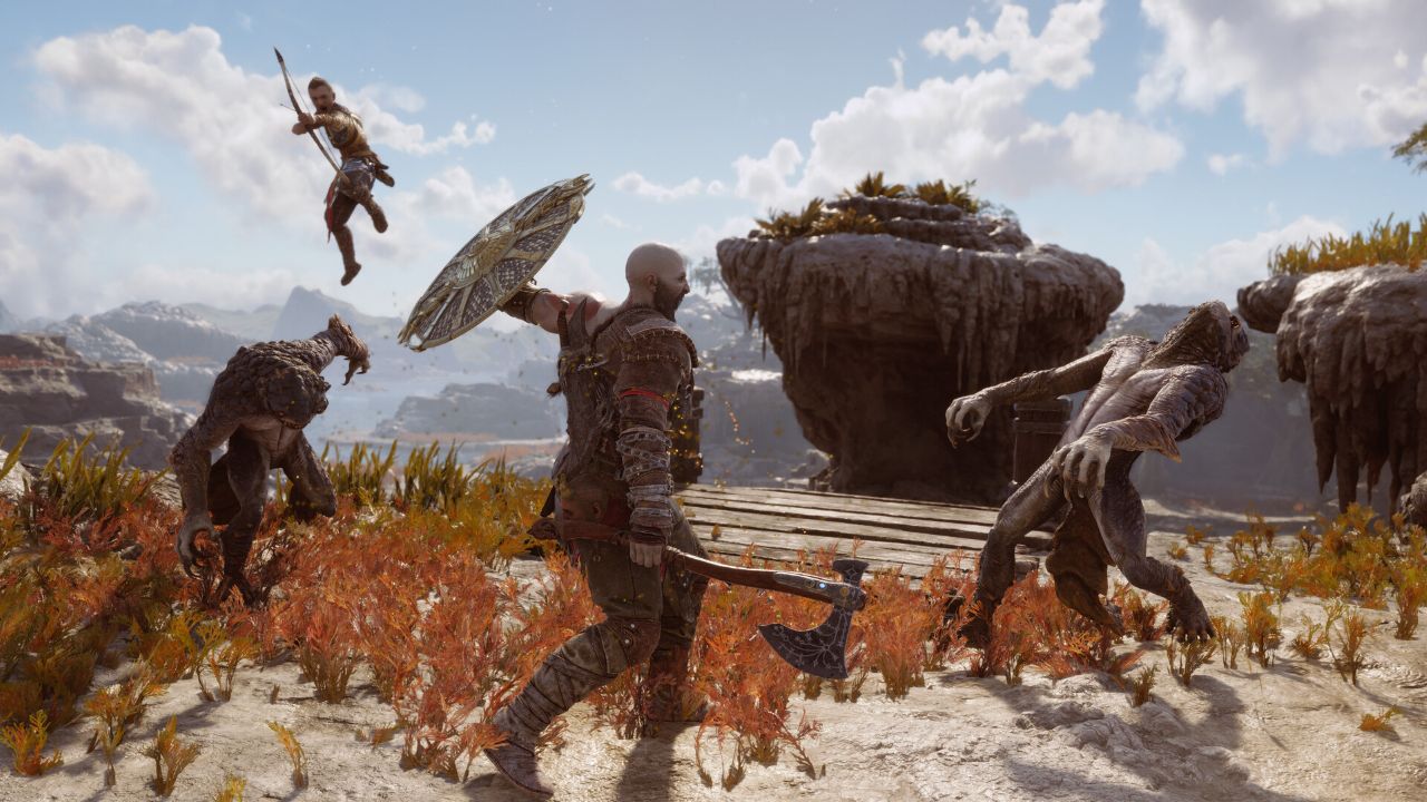 Kratos i Atreus walczą z potworami w otwartym, skalistym krajobrazie z gry God of War Ragnarök. Na środku stoi Kratos – potężnie zbudowany, łysy wojownik z brodą – trzymający w ręce topór bojowy i okrągłą tarczę. Uderza jednego z potworów przypominających humanoidalne bestie o szarej skórze. Po lewej stronie w powietrzu widać Atreusa, który skacze z napiętym łukiem, przygotowany do oddania strzału. Wokół nich znajdują się kolejne potwory atakujące z różnych stron. Scena rozgrywa się na jasnej, kamienistej ziemi porośniętej czerwonawą roślinnością, a w tle widać skały, wodę i jasne niebo z chmurami.