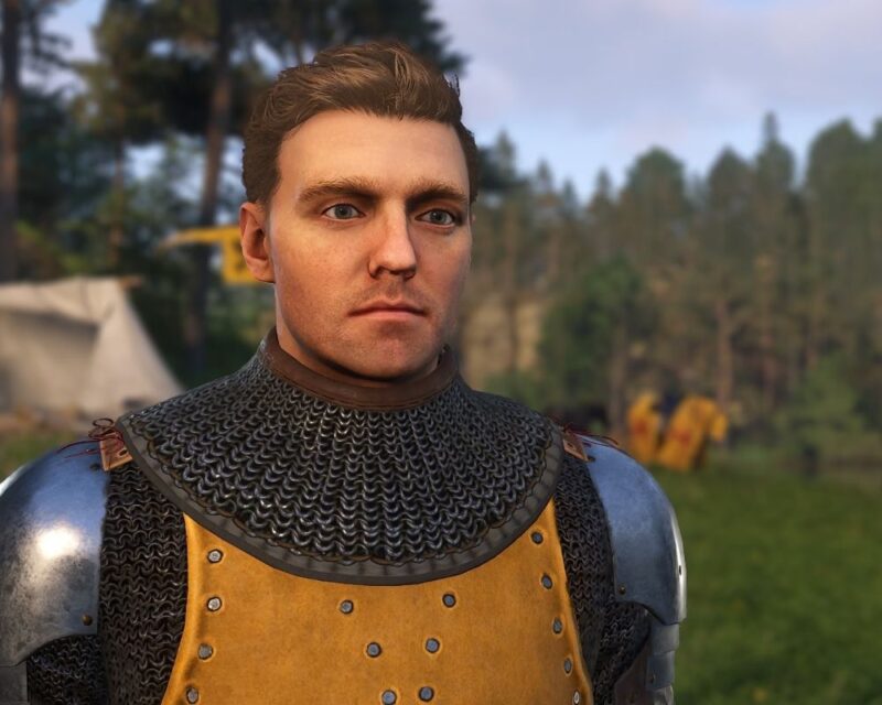 Grafika przedstawia młodego mężczyznę – Henry’ego, głównego bohatera Kingdom Come Deliverance 2, ubranego w średniowieczną zbroję z kolczugą i skórzaną napierśnicą. Stoi na tle leśnego obozu z namiotami i postaciami w oddali.