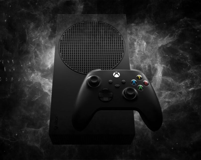 Czarna konsola Xbox Series S Carbon Black z kontrolerem bezprzewodowym na ciemnym tle z efektem dymu. Ilustracja sugeruje, że konsola Xbox Series S przetrwała pożar