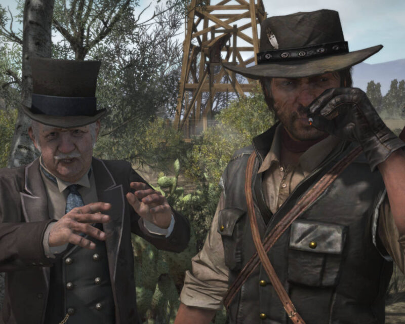Kadr z gry Red Dead Redemption przedstawiający Johna Marstona (po prawej), ubranego w skórzaną kamizelkę, koszulę i kowbojski kapelusz, palącego papierosa i patrzącego w bok. Obok niego stoi starszy mężczyzna w eleganckim, ciemnym garniturze i cylindrze (po lewej), który wykonuje gest uniesionymi dłońmi, jakby coś tłumaczył lub opowiadał. W tle widoczna jest drewniana wieża wiertnicza lub kopalniana oraz pustynna roślinność, w tym kaktusy. Scena sugeruje dialog lub interakcję między tymi dwiema postaciami w świecie Dzikiego Zachodu.