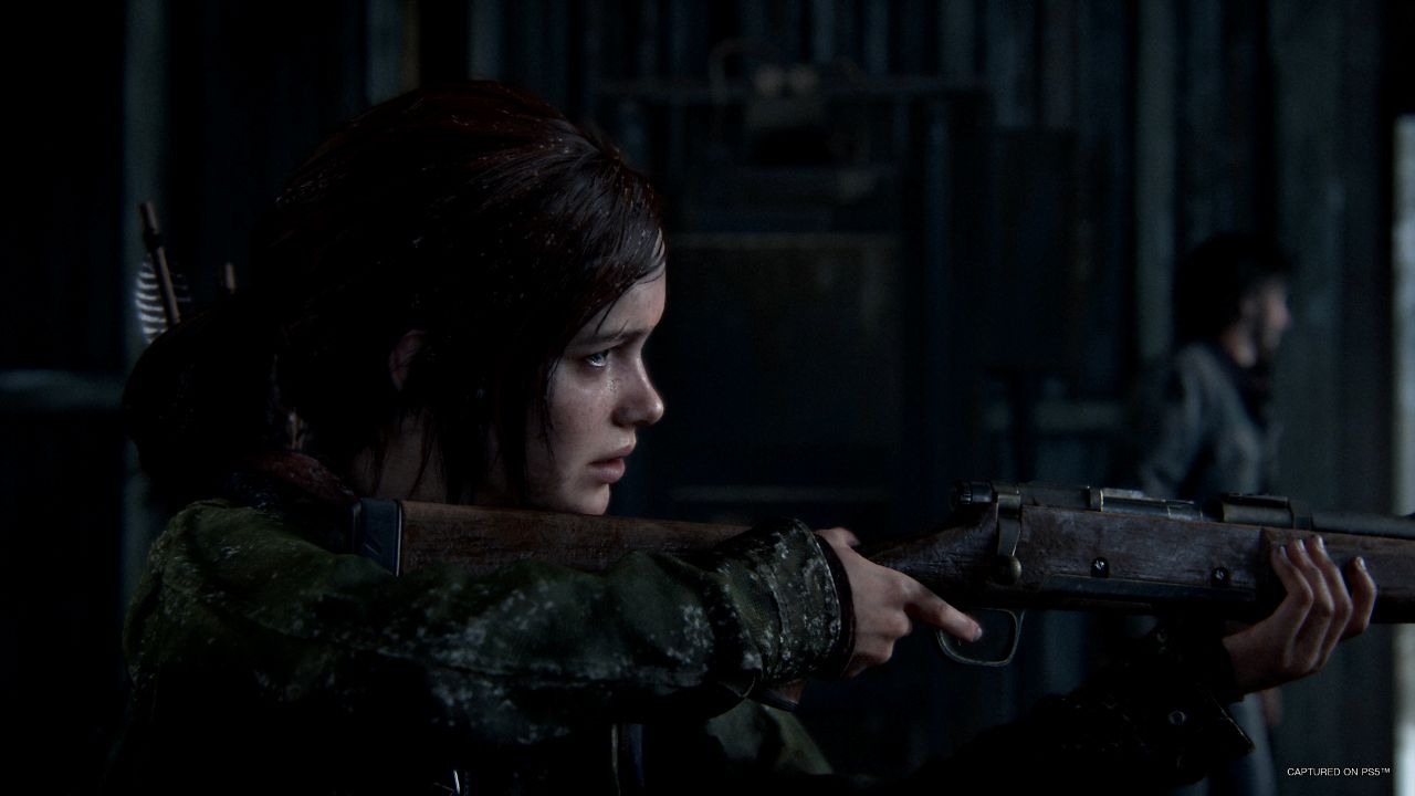 Ellie z gry The Last of Us stoi w ciemnym wnętrzu budynku i celuje z karabinu. Dziewczyna ma brązowe włosy związane z tyłu i ubrana jest w zieloną kurtkę. Trzyma drewniany karabin przy ramieniu, celując przed siebie. Na jej plecach widać łuk i strzały przypięte do plecaka. Scena jest słabo oświetlona, a w tle widać rozmytą sylwetkę innej osoby stojącej w pomieszczeniu z metalowymi ścianami.