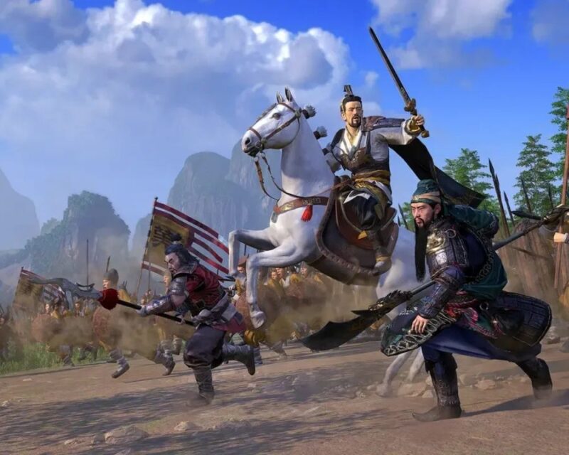 Grafika z gry Total War: Three Kingdoms przedstawia trzech wojowników w tradycyjnych chińskich zbrojach podczas bitwy. Centralna postać na białym koniu unosi miecz, a obok niej biegną dwaj dowódcy z długą bronią drzewcową. W tle widać armię z flagami oraz malownicze, zamglone góry.