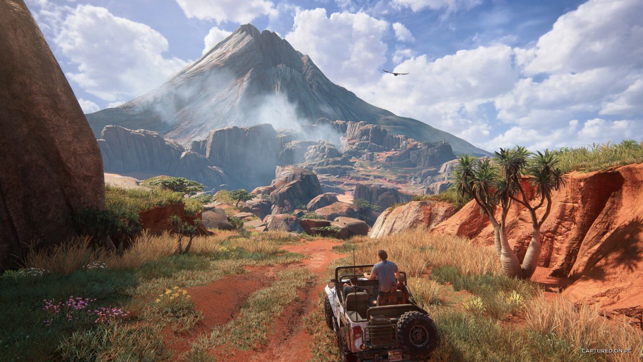 Nathan Drake prowadzi terenowego jeepa jadącego czerwoną, piaszczystą drogą przez tropikalny, górzysty krajobraz z gry Uncharted 4. Obok niego w samochodzie stoi jego brat Sam Drake, trzymając się ramy pojazdu i patrząc w stronę drogi. Auto jedzie przez otwartą przestrzeń porośniętą wysoką trawą, krzewami i kilkoma palmami, a po bokach widać czerwone skały. W tle dominuje ogromna, stroma góra częściowo spowita mgłą, nad którą na tle jasnego nieba z chmurami krąży ptak.