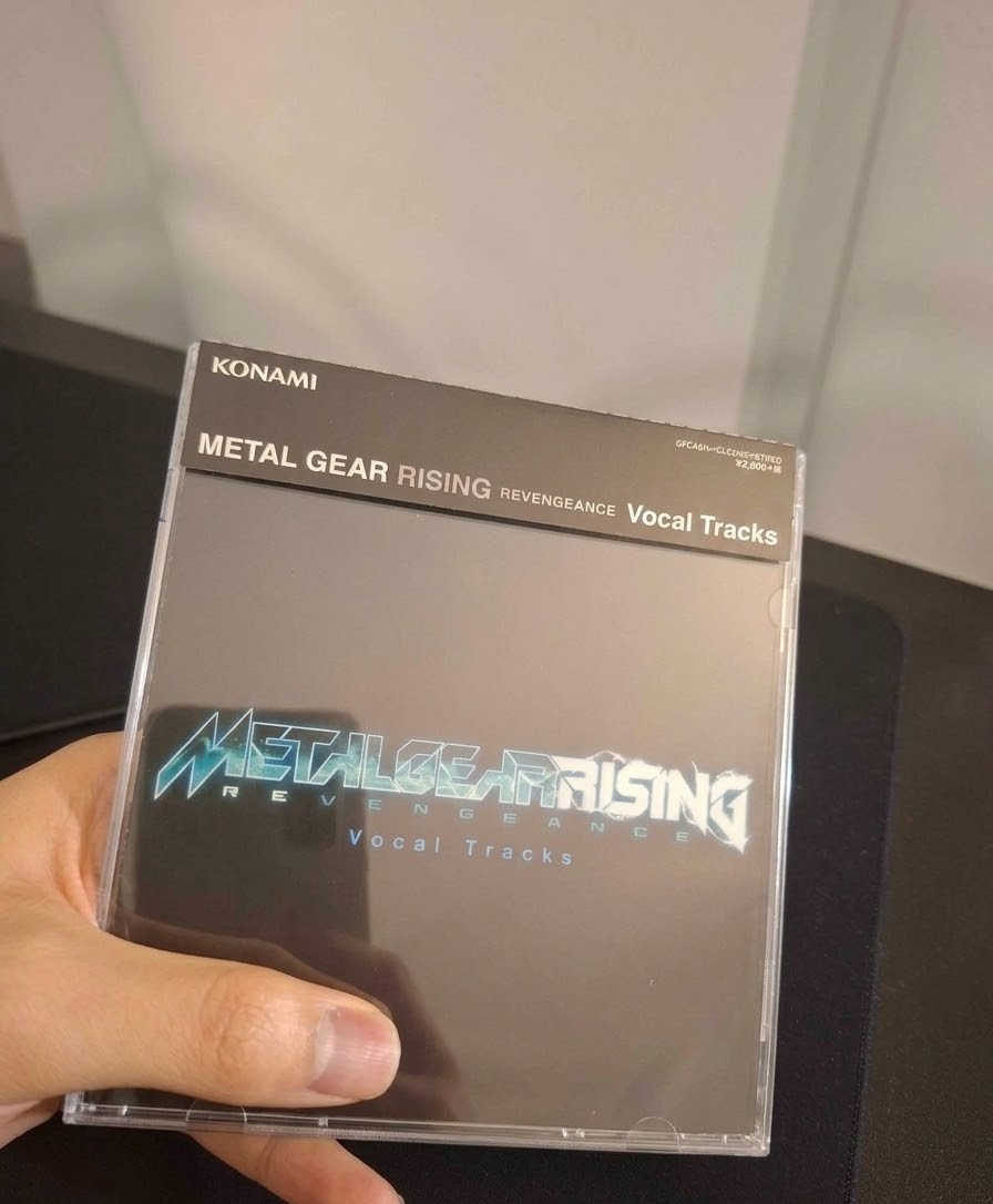 Zdjęcie przedstawia fizyczne wydanie ścieżki dźwiękowej na płycie CD. Opakowanie to klasyczne plastikowe pudełko typu jewel case, na którym znajduje się czarna okładka z charakterystycznym, metalicznym logo gry Metal Gear Rising: Revengeance w odcieniach niebieskiego i bieli. Poniżej głównego logo umieszczono jasnoniebieski napis Vocal Tracks, informujący o zawartości płyty. Górną część pudełka zdobi czarny pasek boczny (obi strip), na którym widnieje logo firmy Konami, pełny tytuł gry oraz cena w jenach, co sugeruje, że jest to wydanie japońskie.