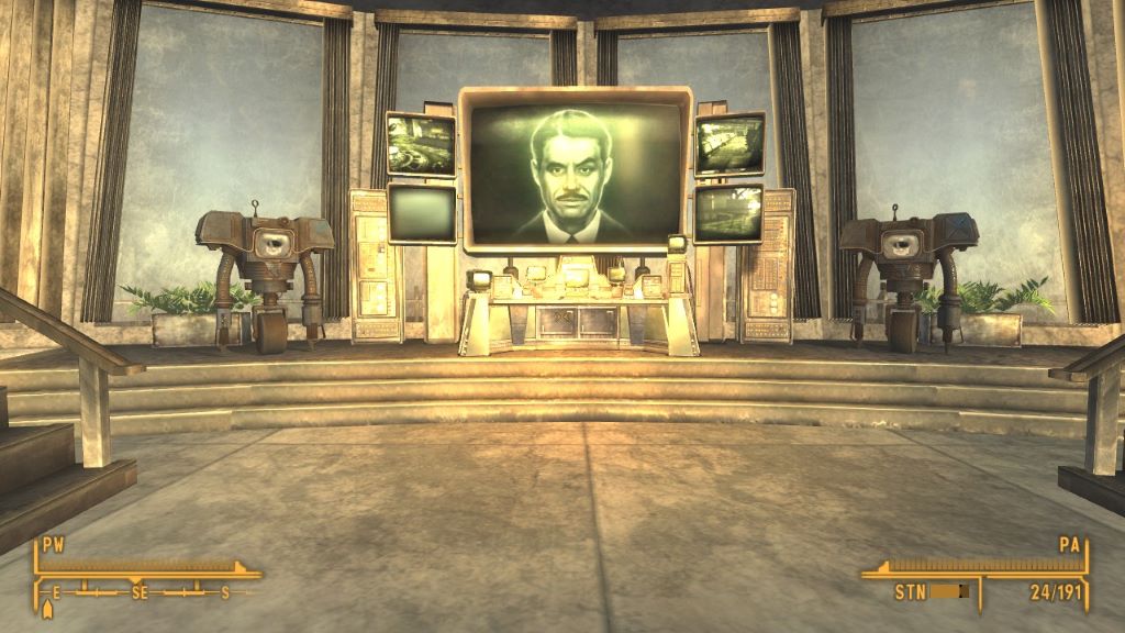 Zrzut ekranu z gry Fallout: New Vegas przedstawiający futurystyczną salę kontrolną o złotawym oświetleniu. W centrum znajduje się duży ekran z twarzą mężczyzny, otoczony mniejszymi monitorami i panelami sterowania. Po obu stronach ekranu stoją symetrycznie dwa humanoidalne roboty na pojedynczych kołach, z pudełkowymi korpusami i mechanicznymi ramionami. W tle widać metaliczne ściany, wysokie okna i elementy retrofuturystycznego wystroju. Na dole ekranu widoczny jest interfejs gracza z kompasem i wskaźnikami statusu, co potwierdza, że to ujęcie z perspektywy pierwszej osoby podczas rozgrywki.