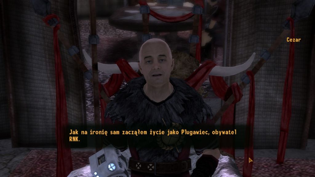 Zrzut ekranu z gry Fallout: New Vegas przedstawiający postać Cezara stojącego na tle czerwonych draperii i dużych, zakrzywionych rogów pełniących funkcję dekoracji. Cezar jest łysy, ubrany w ciemny strój z futrzanym kołnierzem i patrzy prosto w stronę gracza, jakby prowadził rozmowę. Na dole ekranu widoczny jest interfejs dialogowy z polskim napisem: ‘Jak na ironię sam zacząłem życie jako Plugawiec, obywatel RNK.’ Cała scena ma teatralny, ceremonialny charakter, podkreślający władzę i autorytet postaci.