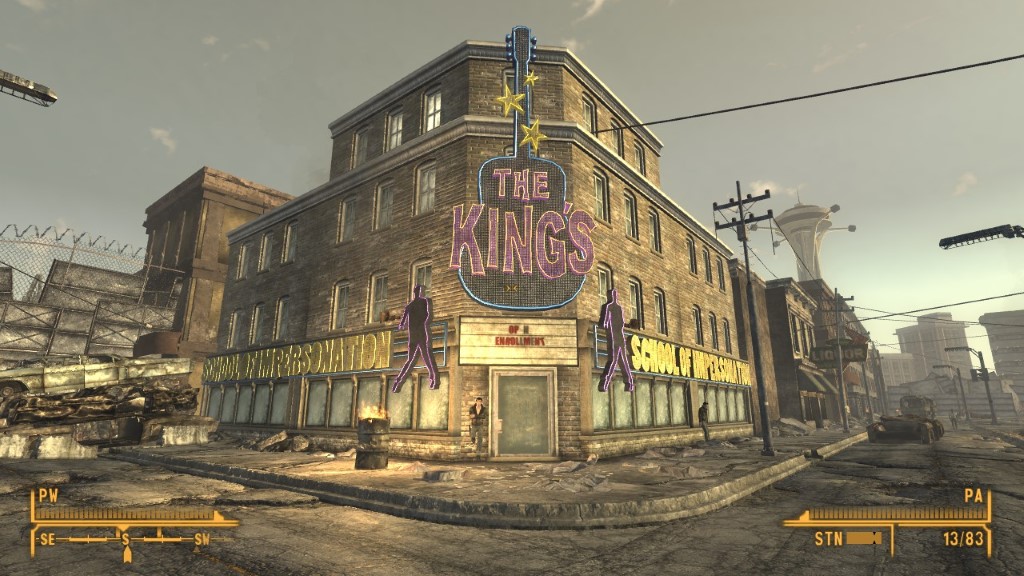 Zrzut ekranu z gry Fallout: New Vegas. W centrum znajduje się duży, wielopiętrowy budynek z cegły, mocno nadgryziony upływem czasu. Na jego fasadzie widnieje ogromny neon w kształcie gitary z napisem ‘THE KING’S’. Poniżej umieszczono kolejny szyld: ‘SCHOOL OF IMPERSONATION’. Ulica przed budynkiem jest zniszczona — widać popękany asfalt, porzucone wraki samochodów i rozsypane gruzowisko. Całość utrzymana jest w postapokaliptycznym klimacie, z przygaszonym, lekko mglistym światłem późnego popołudnia.