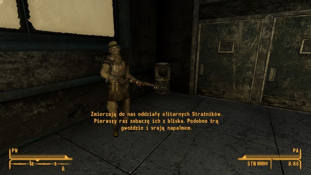 Zrzut ekranu z gry Fallout: New Vegas. W ciemnym, metalicznym pomieszczeniu o industrialnym wystroju stoi postać w ciężkim pancerzu, trzymająca karabin w obu rękach. Ściany i drzwi mają stalowe, nitowane powierzchnie, a oświetlenie jest słabe i chłodne. Na dole ekranu widoczny jest interfejs gracza z kompasem, paskiem zdrowia i innymi wskaźnikami. Na środku ekranu wyświetla się polski tekst dialogowy: ‘Zmierzają do nas oddziały elitarnych Strażników. Pierwszy raz zobaczę ich z bliska. Podobno żrą gwoździe i srają napalmem.’ Cała scena ma surowy, militarno‑postapokaliptyczny charakter.