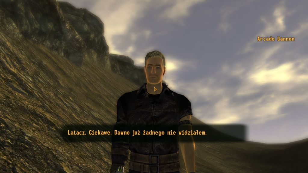Zrzut ekranu z gry Fallout: New Vegas. Na pierwszym planie stoi Arcade Gannon — mężczyzna w ciemnym stroju, patrzący w stronę gracza. Za nim rozciąga się skalisty, pustynny teren z jasnym, częściowo zachmurzonym niebem. Scena ma surowy, naturalny charakter typowy dla pustkowi Mojave. Na dole ekranu widoczny jest interfejs dialogowy z polskim tekstem: ‘Latacz. Ciekawe. Dawno już żadnego nie widziałem.