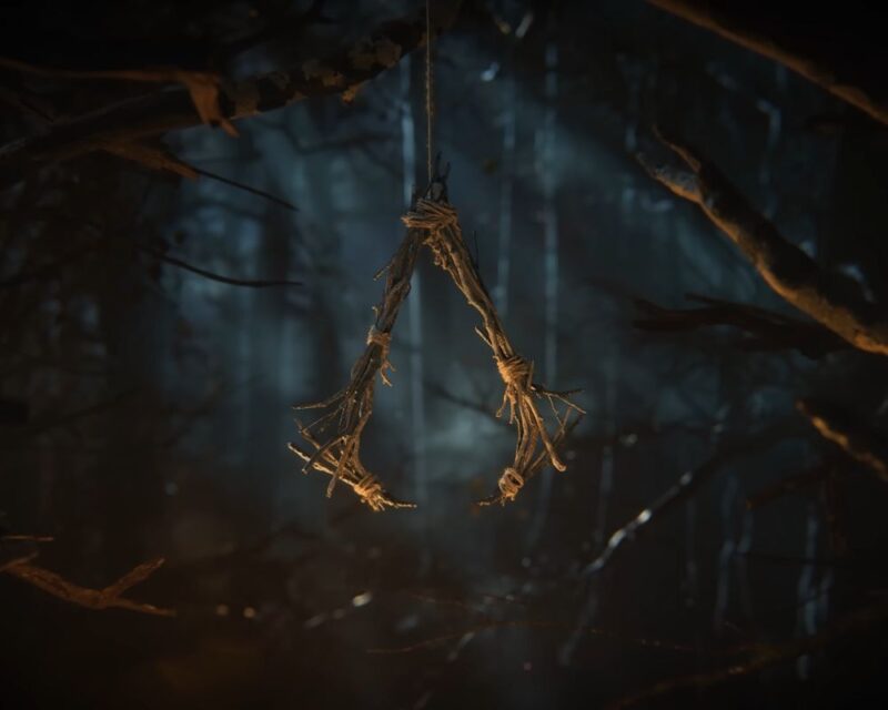 Zrzut ekranu z gry Assassin's Creed Hexe. W centrum widać ikoniczny znak Asasynów, wykonany w formie talizmanu z powiązanych patyków, przypominający estetykę okultystyczną. Symbol wisi na cienkiej lince w gęstym, mrocznym i zamglonym lesie, rozświetlonym jedynie słabym, ciepłym blaskiem z dołu.