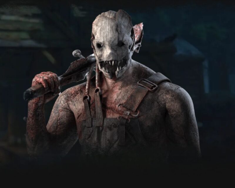 Zrzut ekranu z gry Dead by Daylight przedstawiający postać Trappera (Trapera). Widoczny jest od pasa w górę, ukazując jego potężną sylwetkę pokrytą bliznami i śladami krwi. Postać nosi charakterystyczną, przerażającą maskę zrobioną z kości lub metalu z rzędami ostrych zębów. Na ramieniu opiera swoją broń – zakrwawiony, prowizoryczny tasak. Postać ubrana jest w brudne, ciemne ogrodniczki, a całość utrzymana jest w mrocznej, ponurej kolorystyce.