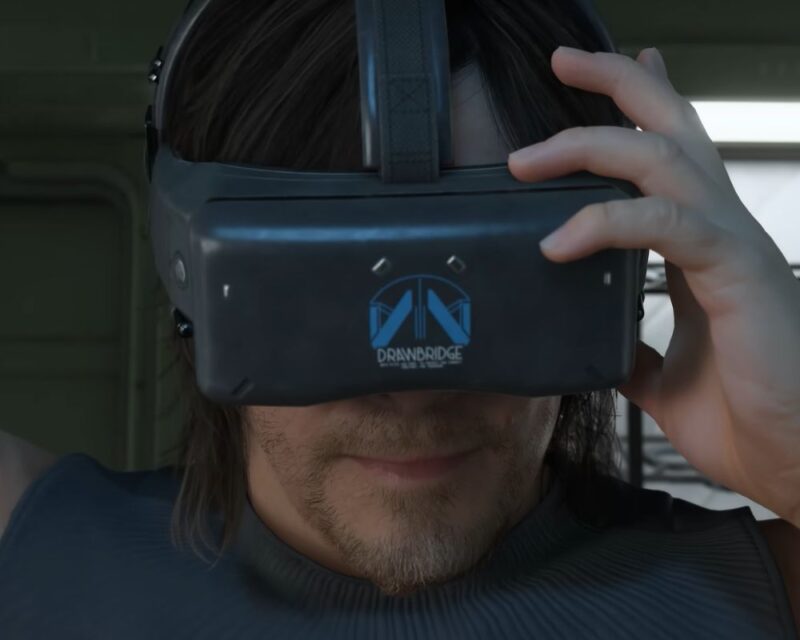 Zbliżenie na Sama Portera Bridgesa zakładającego futurystyczne gogle VR z logo Drawbridge, nawiązujące do gry Death Stranding 2: On the Beach; ujęcie sugeruje immersję w świecie gry, w kontekście wersji na Steam Deck.