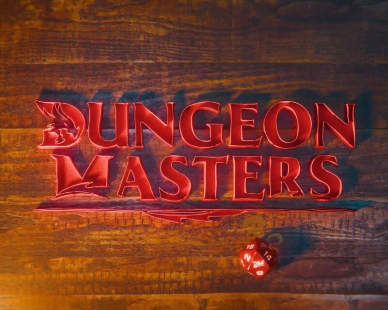 Grafika przedstawia trójwymiarowy, metaliczny logotyp „DUNGEON MASTERS” w kolorze głębokiej czerwieni, umieszczony na tle rustykalnych, drewnianych desek. Stylistyka napisu – w tym charakterystyczne zdobienia liter „D” oraz „M” – bezpośrednio nawiązuje do oficjalnej identyfikacji wizualnej marki Dungeons & Dragons należącej do Wizards of the Coast. Pod napisem widoczna jest czerwona kostka dwudziestościenna (d20), a całość jest oświetlona w sposób podkreślający teksturę drewna i błysk liter.