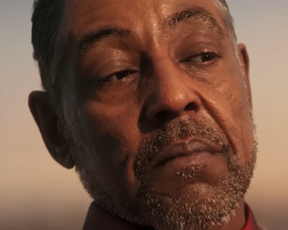 Szczegółowe zbliżenie (portret) na twarz Antóna Castillo, głównego antagonisty z gry Far Cry 6. Mężczyzna ma poważny, zmęczony i surowy wyraz twarzy; jego wzrok jest skierowany nieco w dół i w bok, co nadaje mu melancholijnego, a zarazem groźnego charakteru. Ma krótkie, szpakowate włosy oraz starannie przyciętą brodę i wąsy o wyraźnej, siwej strukturze. Ciepłe, niskie oświetlenie przypominające zachód słońca mocno podkreśla teksturę jego skóry, zmarszczki oraz kontury twarzy, rzucając głębokie cienie. W dolnej części kadru widać kołnierzyk ciemnoczerwonej koszuli, a tło jest miękko rozmyte w odcieniach pomarańczu i beżu.