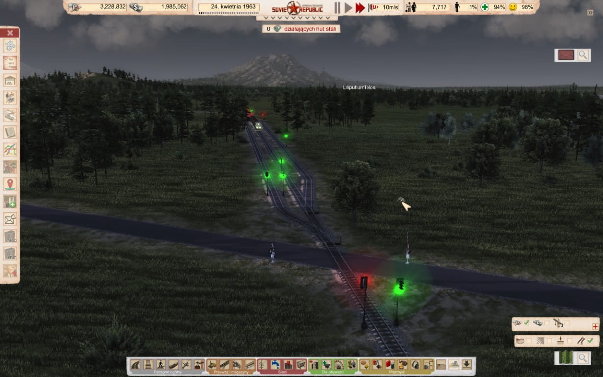 Zrzut ekranu z gry „Workers & Resources: Soviet Republic” przedstawiający skomplikowany węzeł kolejowy o zmierzchu. Na środku widać tory kolejowe z rozjazdami oraz liczne semafory świecące na zielono i czerwono, wyznaczające bloki sygnałowe. Torowisko przecina drogę asfaltową, tworząc przejazd kolejowy. W tle majaczy wysoka, zamglona góra oraz gęsty las.