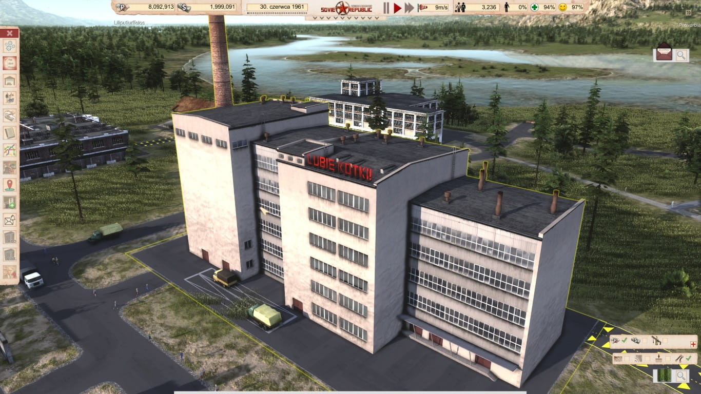 Zrzut ekranu z gry „Workers & Resources: Soviet Republic” przedstawiający duży, wielopiętrowy budynek przemysłowy o jasnej elewacji. Na dachu budynku znajduje się czerwony napis „LUBIĘ KOTKI!!”. Budynek jest zaznaczony żółtą obwódką, a wokół niego widać asfaltowe podjazdy z zaparkowanymi ciężarówkami. W tle rozpościera się krajobraz z rzeką, lasem i wysokim kominem fabrycznym.