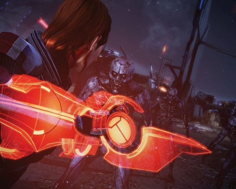 Scena walki z gry Mass Effect – żeńska Shepard, rudowłosa kobieta o krótkich włosach w pancerzu bojowym, walczy przy użyciu czerwonego ostrza energetycznego z grupą zmutowanych przeciwników w zniszczonym, ponurym otoczeniu.