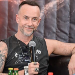 Nergal z zespołu Behemoth. Mężczyzna z tatuażami trzyma mikrofon podczas wywiadu, siedząc na czarnej kanapie. Za nim znajduje się ciemna kurtyna z grafiką w odcieniach czerni i czerwieni.
