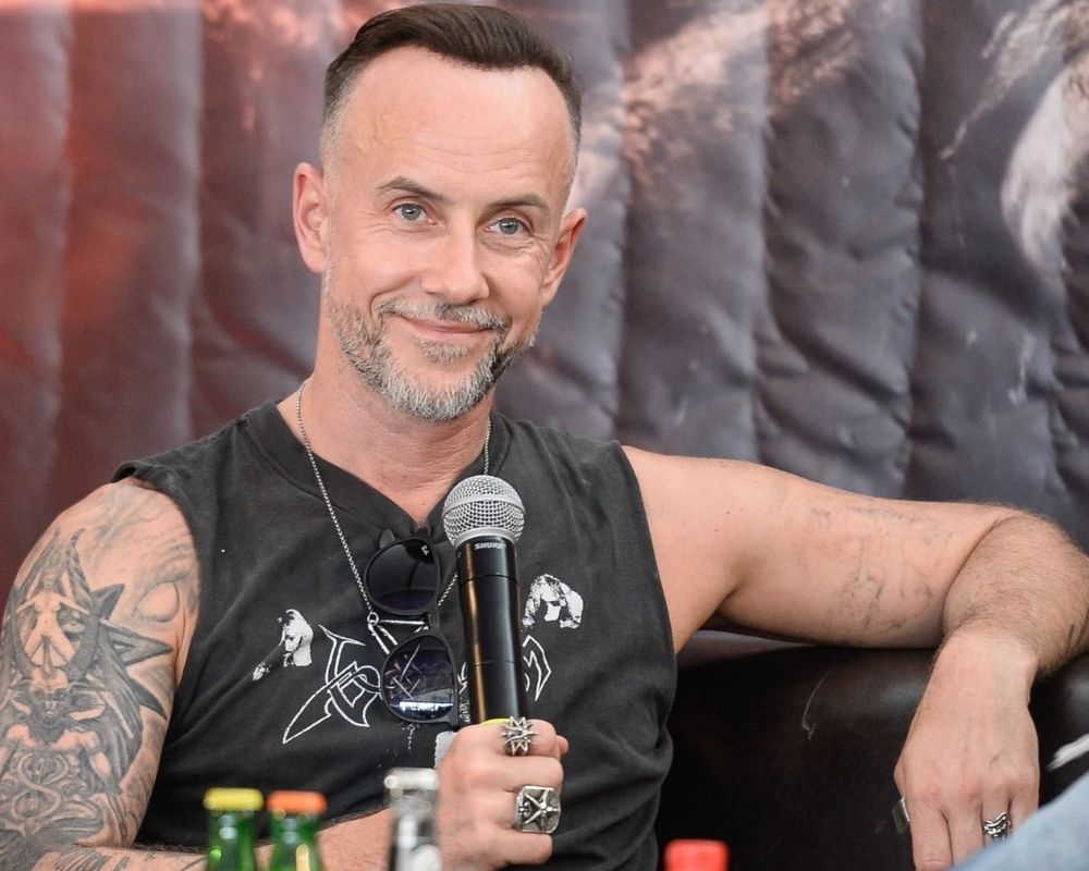 Nergal z zespołu Behemoth. Mężczyzna z tatuażami trzyma mikrofon podczas wywiadu, siedząc na czarnej kanapie. Za nim znajduje się ciemna kurtyna z grafiką w odcieniach czerni i czerwieni.