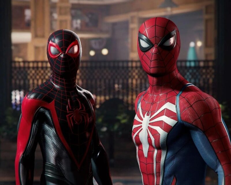 Peter Parker i Miles Morales z gry Spider-Man 2 stoją obok siebie w eleganckim, ciepło oświetlonym wnętrzu przypominającym restaurację lub salę bankietową. Po lewej znajduje się Miles w czarno-czerwonym kostiumie z charakterystycznym czerwonym symbolem pająka i czerwonymi akcentami na masce. Po prawej stoi Peter w klasycznym czerwono-niebieskim stroju z dużym, białym emblematem pająka rozciągającym się na klatce piersiowej. Obaj bohaterowie patrzą w tym samym kierunku, jakby skupieni na czymś poza kadrem.