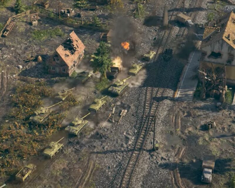 Scena z gry strategicznej Sudden Strike 5 przedstawiająca kolumnę pancerną nacierającą przez zniszczoną wojną wieś, ukazana w rzucie izometrycznym. W centralnej części obrazu kolumna siedmiu oliwkowozielonych czołgów T-34 przedziera się przez głębokie, wypełnione wodą błoto i koleiny. W samym środku formacji jeden z pojazdów został trafiony – z jego wieży bucha jaskrawopomarańczowy ogień, a w górę unosi się potężny, gęsty słup czarnego dymu, który przesłania fragmenty otoczenia. Po lewej stronie drogi stoi zniszczony dom z czerwonej cegły z dużą dziurą w dachu, obok którego widać drewnianą studnię, płoty oraz liczne skrzynie z zaopatrzeniem. Po prawej stronie, za podwójną linią zardzewiałych torów kolejowych, znajduje się uszkodzony, jasny budynek oraz całkowicie zrujnowana, drewniana konstrukcja przypominająca wiatrak. W tle i na poboczach widoczne są trzy wojskowe ciężarówki oraz rozrzucone beczki. Całość sceny skąpana jest w surowych, przygaszonych barwach ziemi, rdzy i militarnej zieleni, co podkreśla realistyczny i ponury klimat pola bitwy.