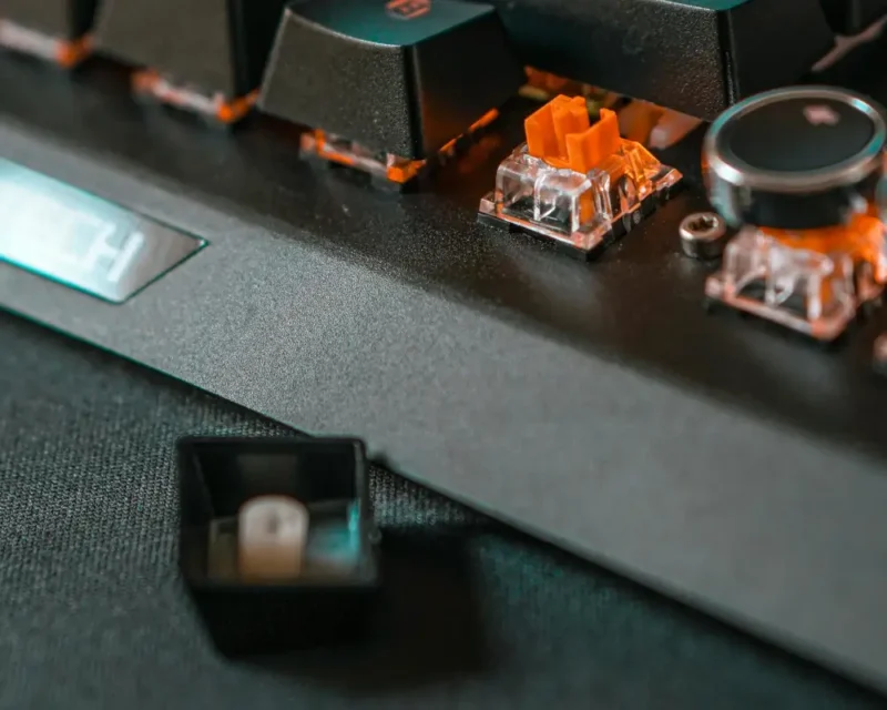Zdjęcie przedstawia artystyczne zbliżenie na mechaniczną klawiaturę gamingową o matowym, czarnym wykończeniu, na której wyeksponowano pomarańczowy przełącznik (switch) po zdjęciu jednego z klawiszy. Odpięty czarny kapsel leży na ciemnej, teksturowej podkładce na pierwszym planie, ukazując swoje wewnętrzne mocowanie, podczas gdy w tle widać rzędy pozostałych klawiszy z delikatnym, kolorowym podświetleniem. Po prawej stronie kadru znajduje się metalowe pokrętło głośności oraz fragmenty obudowy z widoczną śrubą montażową, a po lewej stronie majaczy podświetlony logotyp marki, co nadaje całości nowoczesny, technologiczny klimat podkreślony przez niską głębię ostrości i ciepłe, pomarańczowe akcenty świetlne wydobywające się spod przycisków.