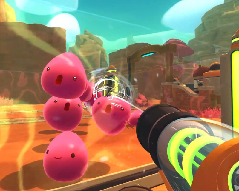 Na obrazku widać screen z gry Slime Rancher. Przedstawia kolorową, bajkową scenę z pustynnym krajobrazem. W tle znajdują się skały i futurystyczne konstrukcje. Na pierwszym planie widzimy kilka różowych slime'ów, które podskakują. Z prawej strony obrazka widać vackpack, czyli urządzenie odpowiedzialne za zbieranie rzeczy w grze.