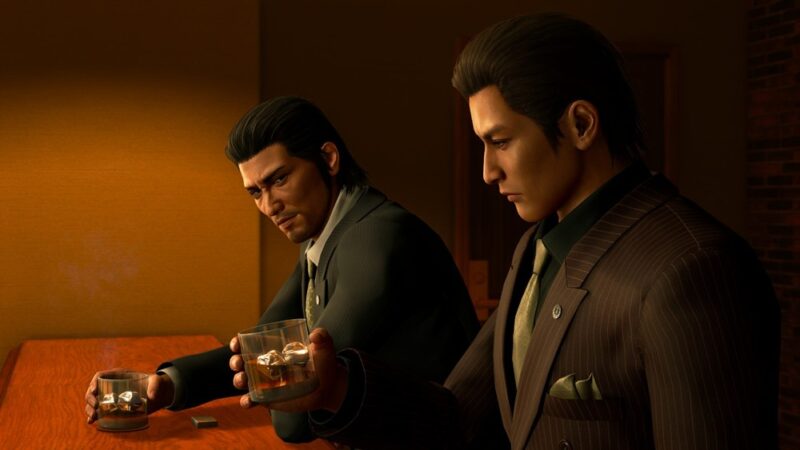 Scena z gry wideo Yakuza Kiwami 3 & Dark Ties, gdzie dwóch mężczyzn w garniturach siedzi przy barze i piją whisky ze szklanek. Otoczenie jest drewniane i słabo oświetlone