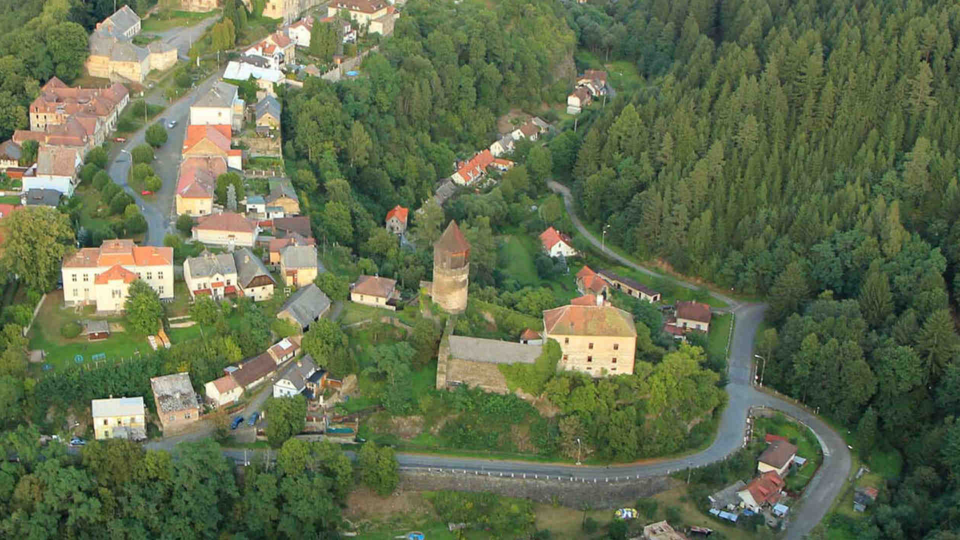 Na obrazku widzimy zielone wzgórza i lasy. Po lewej stronie pośród drzew widać dachy domów. Domy są głównie białe z brązowmi dachami, Na środku znajduje się średniowieczny zamek Pirkstejn. Jego najbardziej charakterystyczną cechą jest wieża o okrągłym kształcie.