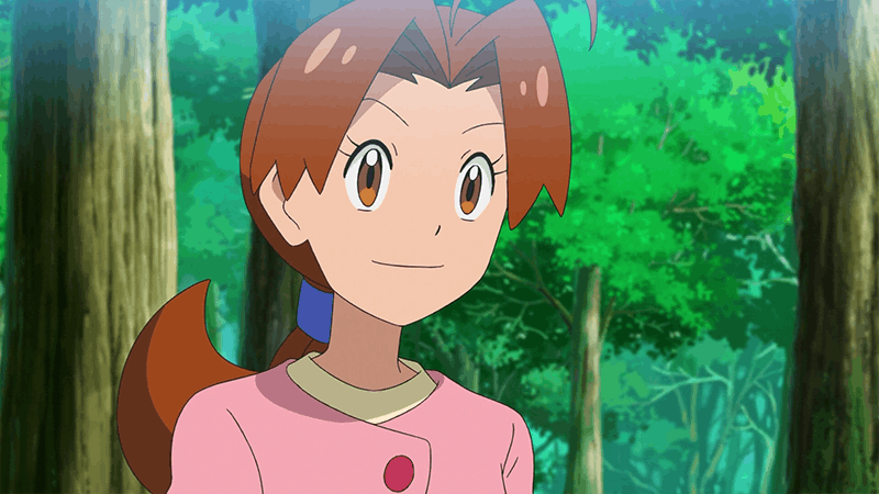 delia pokemon Kadr z anime przedstawiający Delię Ketchum z serii Pokémon. Młoda kobieta o jasnej karnacji, brązowych oczach i kasztanowych włosach spiętych z tyłu niebieską gumką patrzy lekko w bok z łagodnym uśmiechem. Ma na sobie charakterystyczną różową bluzkę z kremowym kołnierzykiem i dużym, okrągłym guzikiem. Tło stanowi rozmyty, zielony las z widocznymi pniami drzew.