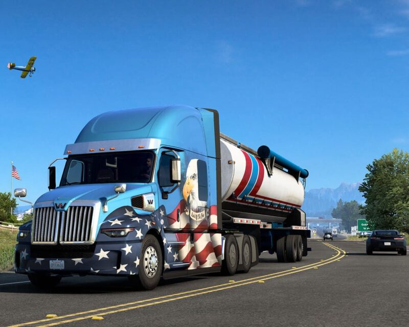Grafika pochodzi z gry American Truck Simulator. Na pierwszym planie widzimy niebiesko-białą ciężarównkę. Z przodu samochodu znajduje się grafika z motywem orła na amerykańskiej fladze. Ciężarówka jedzie po drodze. W tle widzimy lecący samolot.