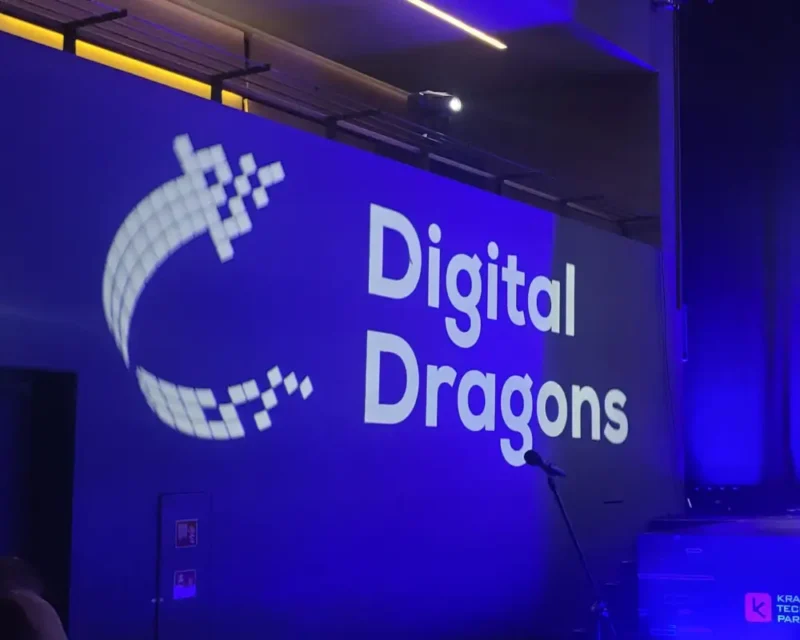 Na dużej, ciemnej ścianie (lub ekranie) wyświetlany jest biały napis „Digital Dragons” (część nazwy słynnej konferencji branży gier wideo). Po lewej stronie tekstu znajduje się charakterystyczne logo w kształcie stylizowanej, otwartej litery „C” lub smoczego ogona, złożonego z pikselowych, białych kwadracików.