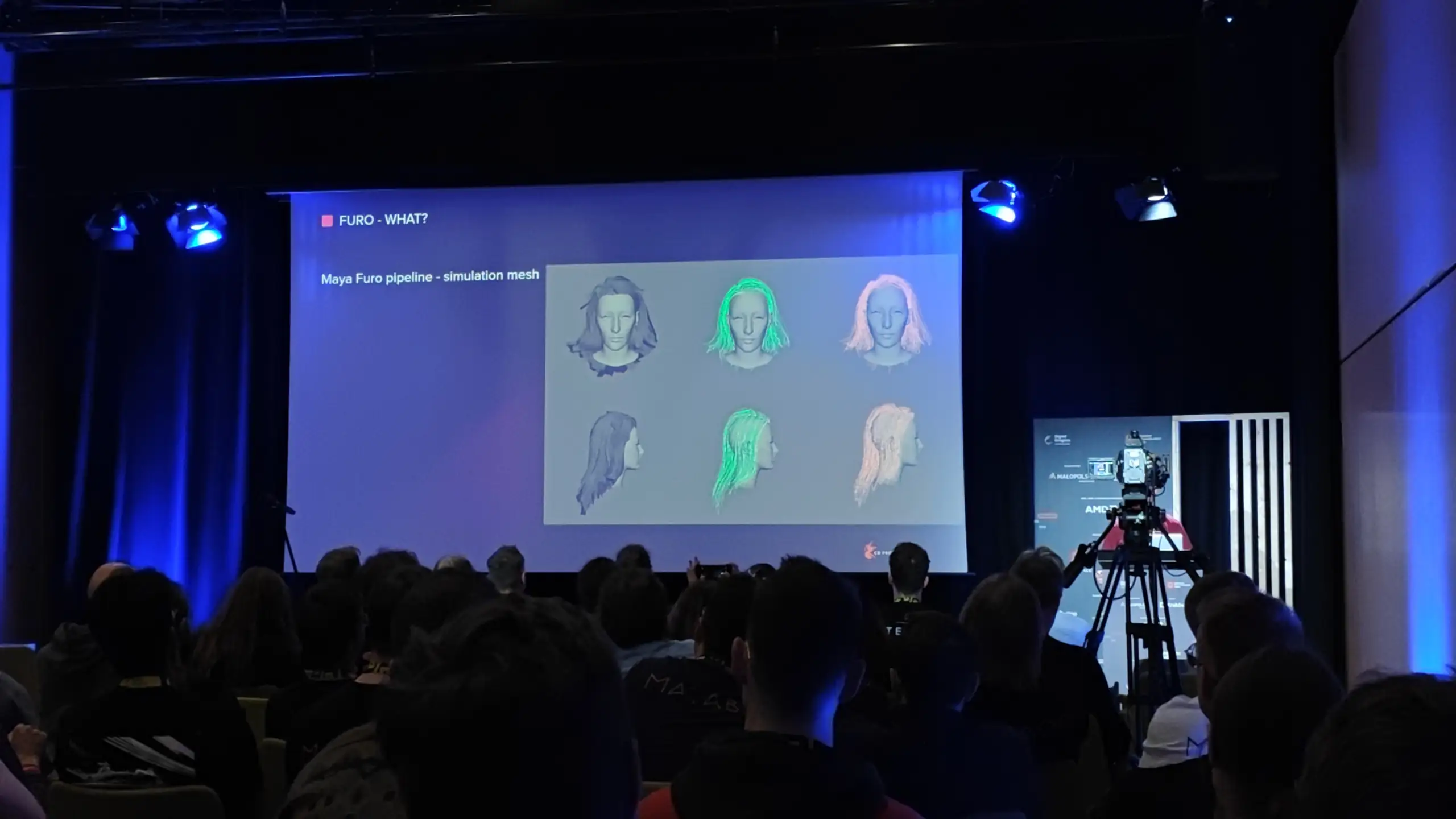 Zdjęcie przedstawia prelekcję odbywającą się w zaciemnionej sali konferencyjnej, gdzie na dużym ekranie w centrum wyświetlana jest techniczna prezentacja zatytułowana „FURO - WHAT?” oraz „Maya Furo pipeline - simulation mesh”, ukazująca sześć trójwymiarowych modeli ludzkich głów z różnymi etapami symulacji włosów (od szarych siatek po wersje podświetlone na zielono i różowo). Scena i boki sali są oświetlone niebieskim światłem reflektorów, a w prawym rogu, tuż obok ekranu, na statywie stoi profesjonalna kamera telewizyjna skierowana w stronę sceny. Na pierwszym planie oraz w środkowej części kadru widoczne są liczne sylwetki tyłem siedzącej publiczności, która w skupieniu ogląda prezentację, tworząc atmosferę branżowego wykładu technologicznego związanego z grafiką komputerową lub tworzeniem gier wideo.