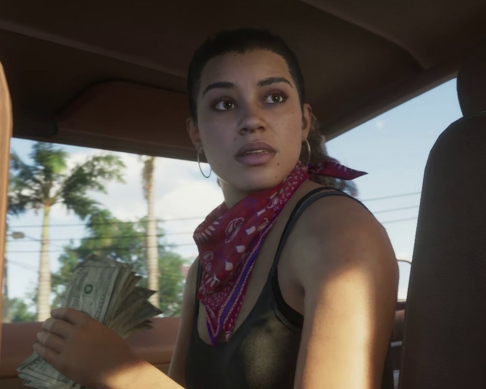 Lucia z GTA 6 siedzi we wnętrzu samochodu i trzyma w lewej dłoni gruby plik dolarów. Bohaterka ma ciemne włosy związane z tyłu, duże kolczyki koła oraz czerwoną bandanę zawiązaną na szyi. Ubrana jest w czarny top, a jej twarz oświetla ciepłe światło wpadające przez okno auta. Lucia spogląda nerwowo za siebie, jakby reagowała na coś dziejącego się poza kadrem. W tle widoczne są palmy, przewody elektryczne i słoneczne niebo.