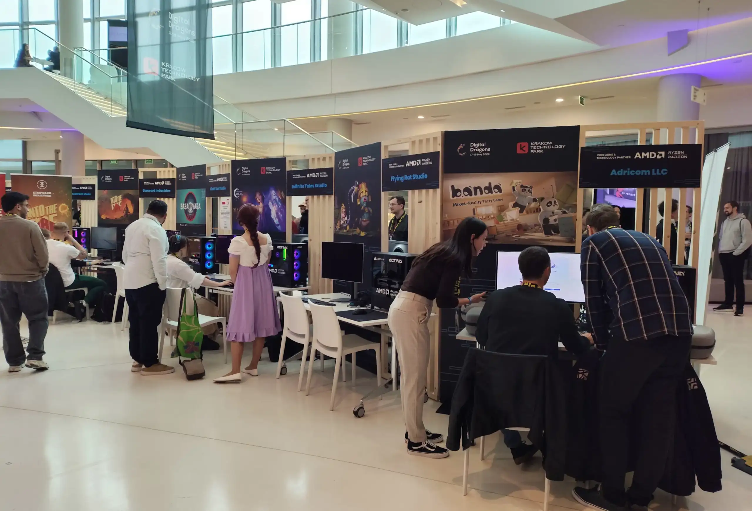 Zdjęcie przedstawia przestronną, nowoczesną halę wystawową podczas wydarzenia „Digital Dragons”, gdzie zorganizowano strefę gier niezależnych (Indie Zone) z rzędami drewnianych boksów i stanowisk komputerowych. Liczni odwiedzający oraz deweloperzy gromadzą się wokół monitorów, testując gry wideo, rozmawiając lub obserwując rozgrywkę. Na pierwszym planie po prawej stronie widoczne jest stanowisko gry „Banda” (Mixed-Reality Party Game), przy którym stoi kobieta w jasnych spodniach i ciemnej bluzce oraz mężczyzna w kraciastej koszuli, przyglądający się osobie siedzącej przed ekranem komputera. Po lewej stronie uwagi doświadcza inne stanowisko, gdzie stoi kobieta w fioletowej spódnicy oraz mężczyzna w białej koszuli, a obok nich inni uczestnicy testują przygotowane produkcje. Nad boksami wiszą czarne banery z logotypami „Digital Dragons”, „Krakow Technology Park” oraz partnerów technologicznych, takich jak AMD Ryzen i Radeon, a w tle widać wielopoziomową architekturę budynku z przeszklonymi balustradami, schodami i dużymi oknami wpuszczającymi naturalne światło.