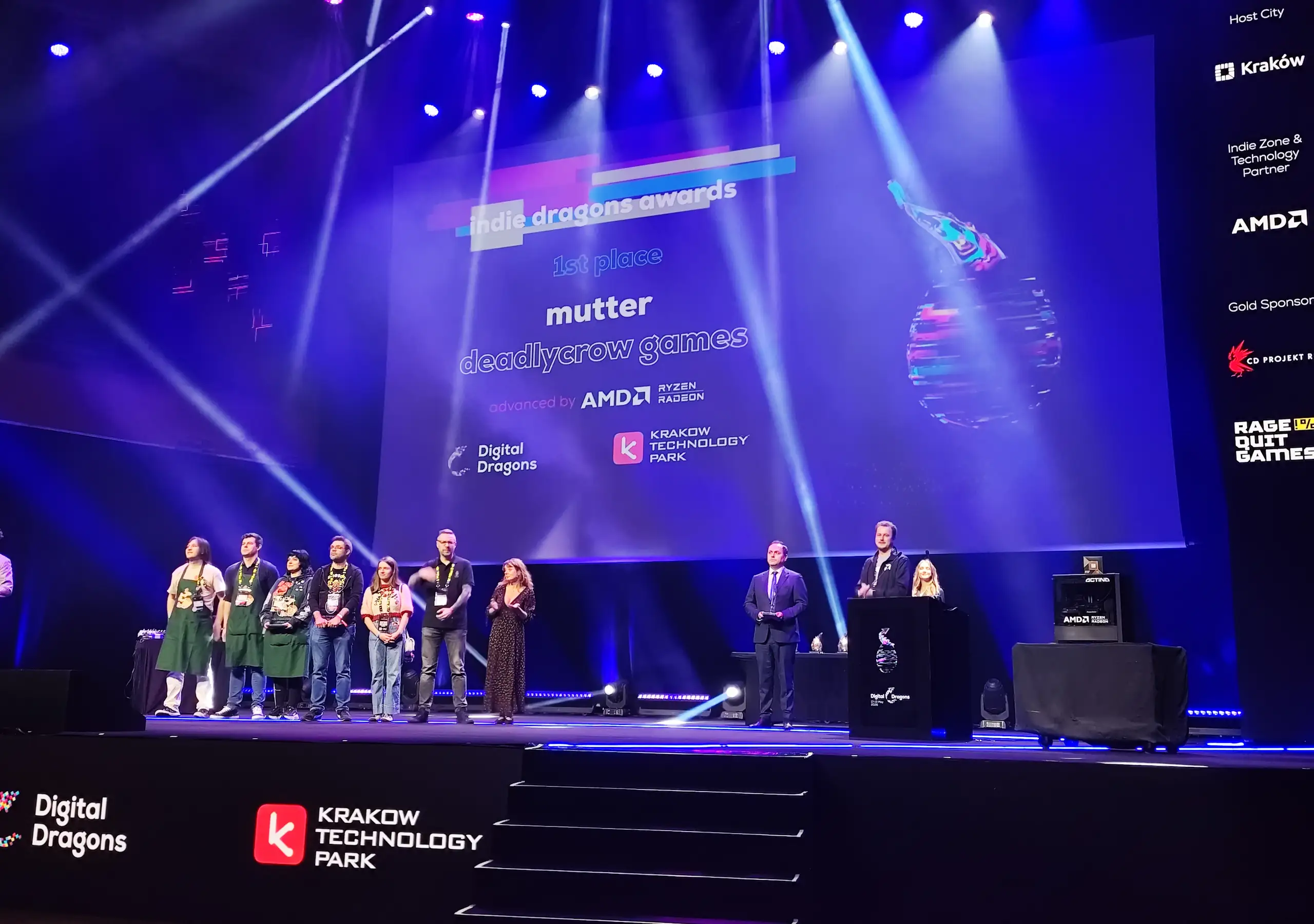 Zdjęcie przedstawia uroczystą galę wręczenia nagród „Indie Dragons Awards” na dużej scenie oświetlonej mocnymi, niebieskimi snopami reflektorów. W centralnej części tła dominuje ogromny ekran multimedialny wyświetlający informację o zdobywcy pierwszego miejsca (1st place) – grze o tytule „Mutter” stworzonej przez „Deadlycrow Games”, z widocznymi logotypami partnerów: AMD Ryzen Radeon, Digital Dragons oraz Krakow Technology Park. Po prawej stronie ekranu widać grafikę przedstawiającą postać nurkującą w kolorową, glitchową kulę. Na scenie stoi grupa ludzi: po lewej stronie ustawiona jest linia laureatów lub nominowanych w różnorodnych strojach (od zielonych fartuchów po luźne koszulki i jeansy), w centrum stoi mężczyzna w eleganckim garniturze, a po prawej stronie, za czarną mównicą z logo Digital Dragons, znajdują się prowadzący wydarzenie. Na samym skraju po prawej stronie sceny umieszczono stanowisko komputerowe, po prawej stronie nad ekranem widoczne są loga sponsorów (m.in. CD Projekt Red, AMD), a u dołu konstrukcji sceny, przy schodkach, wyraźnie odznaczają się podświetlone napisy Digital Dragons oraz Krakow Technology Park.