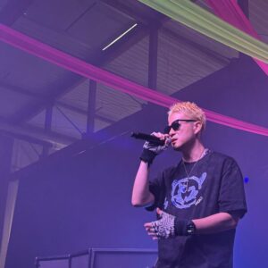 Na zdjęciu widzimy rapera Lotus Juice'a. Mężczyzna ma blond włosy. Ma na sobie okulary przeciwsłoneczne, luźny, czarny t-shirt, czarną bandanę na nadgarstku oraz biało-czarne rękawiczki. W prawej ręce trzyma mikrofon. W tle widzimy halę, w której odbywał się koncert. Przyozdobiono ją kolorowymi szarfami (różowe i zielone) oraz światłami (tutaj fioletowo-niebieskie).